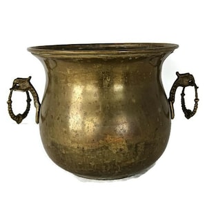 May include: A vintage brass planter with a wide, rounded body and two ornate handles. The handles are attached to the sides of the planter and feature a decorative design. The planter has a slightly flared rim and a weathered appearance.