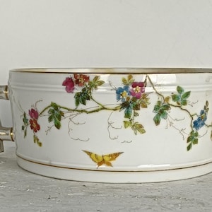 May include: A white ceramic planter with a floral design featuring pink, blue, and yellow flowers. The planter has two decorative handles and a gold trim. A yellow butterfly is painted on the front.