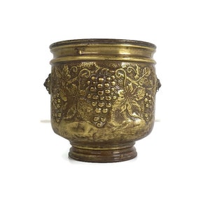 Könnte beinhalten: Ein goldfarbener Metalltopf mit einem Weinreben-Design. Der Topf hat eine runde Form und einen breiten Boden.