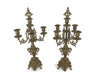 May include: Two antique brass candelabras with ornate detailing. Each candelabra has five candle holders and a decorative base. The brass has a warm, aged patina, and the design is symmetrical and elegant. The candelabras are on a white background.