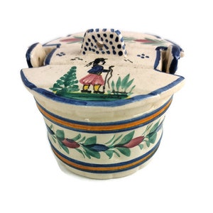 Puede incluir: Un recipiente de mantequilla de cerámica blanca con tapa. La tapa tiene un asa de lunares azules y blancos y una escena pintada de una persona con un vestido azul y rojo caminando con un palo. El recipiente tiene una franja azul y naranja alrededor de la parte superior y un diseño floral alrededor de la parte inferior.