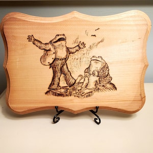 Può includere: Targa in legno con un'illustrazione pirografata di due rane antropomorfe. Una rana è in piedi con le braccia tese, indossa un abito. L'altra rana è seduta. La targa ha un bordo decorativo ed è esposta su un supporto in metallo nero.