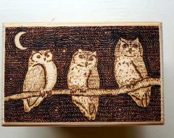 Op de afbeelding: Een houten doos met een gebrandschilderd ontwerp van drie uilen die op een tak zitten. De uilen zijn bruin en wit met grote ogen. De achtergrond is donkerbruin met een halve maan.