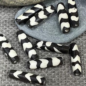 May include: A collection of black and white patterned beads. The beads are cylindrical with a repeating chevron design in black and white. The beads are made of a dark material and have a smooth finish. The beads are for jewelry making.