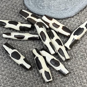 May include: A collection of wooden beads with a black and white cow print pattern. The beads are cylindrical and vary in length. A gray stone rests in the background.
