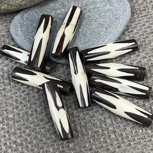 May include: A collection of handcrafted, cylindrical beads. Each bead is dark brown with a white, zigzag pattern. The beads are scattered on a textured, gray surface, suggesting a natural or rustic aesthetic. These beads could be used for jewelry making or other craft projects.