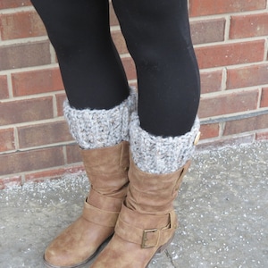 May include: A pair of brown suede boots with a buckle detail, paired with black leggings and gray and white knit leg warmers. The leg warmers have a button detail on the side. The boots are approximately 10 inches tall.