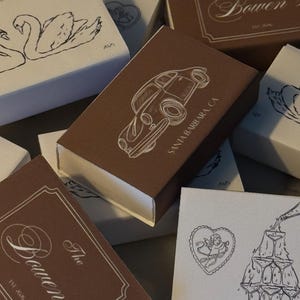May include: A collection of rectangular matchboxes in brown and white featuring various line art illustrations including a vintage car with the text SANTA BARBARA CA, a champagne tower, swans, and the text The Bowen's EST 2026 in a decorative frame.
