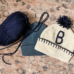 Op de afbeelding: Twee gebreide mutsen, een beige met een zwarte pompon en de letter "B" erop geborduurd, en een donkerblauwe met een zwarte bol wol.