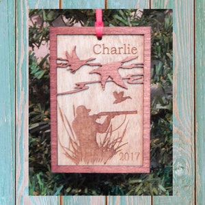 May include: Wooden ornament with a hunting scene, featuring a hunter aiming a rifle, ducks in flight, and the name "Charlie" above the year "2017". The ornament is framed in a darker wood and has a red ribbon for hanging.