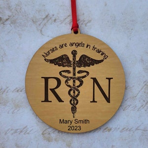 May include: A round wooden ornament with a red ribbon for hanging. The ornament features the text "Nurses are angels in training" and "RN" with a medical symbol. The name "Mary Smith" and the year "2023" are also engraved.