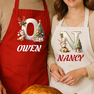 Puede incluir: Dos delantales navideños, uno rojo y otro blanco. El delantal rojo presenta el nombre "OWEN" y la letra "O" con un árbol de Navidad y un reno. El delantal blanco muestra "NANCY" y la letra "N" con decoraciones festivas.