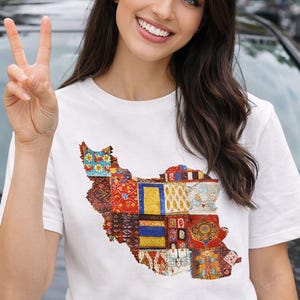 May include: White t-shirt featuring a colorful, patchwork design in the shape of a country. The design incorporates various patterns and colors, including red, blue, yellow, and gold. The shirt is short-sleeved.