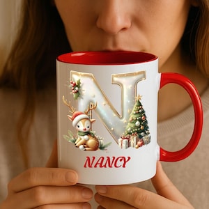 May include: White ceramic mug with a red interior and handle. The mug features a gold-outlined letter "N" with a Christmas tree, gifts, and a reindeer wearing a Santa hat. The name "Nancy" is printed in red.