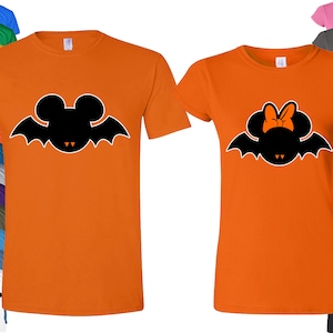 Puede incluir: Dos camisetas naranjas con una silueta negra de Mickey Mouse y Minnie Mouse vestidos de murci&eacute;lagos. Mickey Mouse tiene colmillos y Minnie Mouse tiene un lazo.