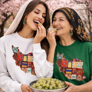 May include: Two people wearing white and green shirts with a colorful map of Iran design. The white shirt is a long-sleeve sweatshirt. The green shirt is a short-sleeve t-shirt. Both are smiling and eating grapes.