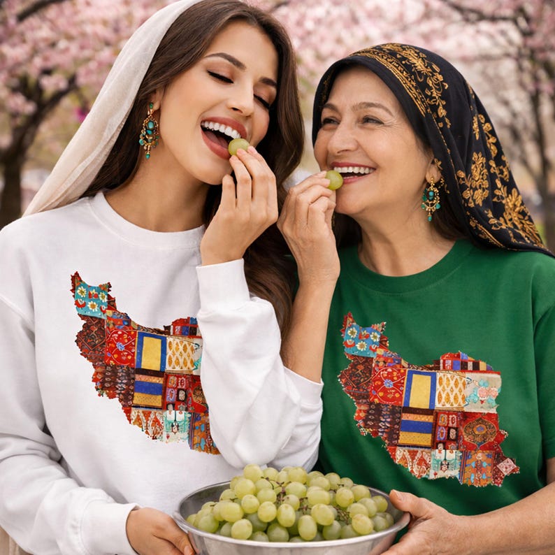 May include: Two people wearing white and green shirts with a colorful map of Iran design. The white shirt is a long-sleeve sweatshirt. The green shirt is a short-sleeve t-shirt. Both are smiling and eating grapes.