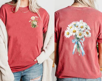 Op de afbeelding: Een baksteenrood t-shirt met korte mouwen, voorzien van een kleine bos witte madeliefjes op de linkerborst en een grote bos witte madeliefjes vastgebonden met een lichtblauw lint op de achterkant van het kledingstuk.