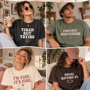 May include: A collection of four short-sleeve t-shirts featuring graphic text. One charcoal shirt reads TIRED BUT TRYING, one olive shirt reads PROBABLY OVERTHINKING, one cream shirt reads I'M FINE. IT'S FINE., and one dark brown shirt reads SOCIAL BATTERY: 2%.