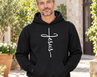 Puede incluir: Una sudadera con capucha negra que presenta un diseño gráfico blanco de una cruz donde la línea vertical está formada por la palabra Jesus escrita en letra cursiva. La prenda incluye un bolsillo tipo canguro y mangas largas.