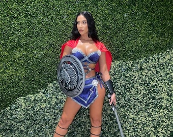 May include: A woman dressed as a warrior, wearing a blue and silver costume with a red cape, holding a shield and a sword. She is standing in front of a green wall.