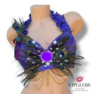 Puede incluir: Un top halter morado con plumas de pavo real, pedrer&iacute;a y una gran gema morada. Cadenas doradas con perlas cuelgan de la parte inferior. El top est&aacute; en un maniqu&iacute;. El logotipo de Lipgloss Costume est&aacute; en la esquina inferior derecha.