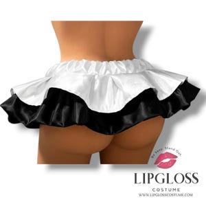 K&ouml;nnte beinhalten: Ein zweilagiger Minirock aus schwarzem und wei&szlig;em Satin mit R&uuml;schen. Der Rock hat eine wei&szlig;e obere Schicht und eine schwarze untere Schicht, mit einem elastischen Bund. Das Bild enth&auml;lt den Text "LIPGLOSS COSTUME".