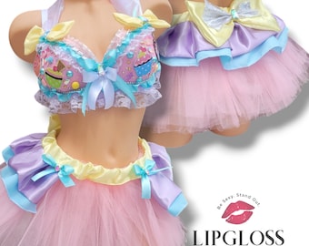May include: A pink tulle skirt with a yellow satin waistband and a lavender satin overlay. The skirt has a blue satin bow on the side. The skirt is paired with a pink bra with a white lace overlay and a cupcake design. The bra has a yellow satin bow on the front. The bra and skirt are part of a costume set.