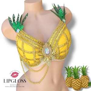 Pode incluir: Um sutiã amarelo com tema de abacaxi com detalhes dourados e uma peça central de joias. O sutiã tem folhas verdes nas alças. O texto "Lipgloss Costume" é visível na parte inferior da imagem.