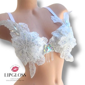 May include: White, embellished costume top with floral and leaf-like designs. The top is covered in sequins and iridescent gems, with dangling bead accents. The straps are white and the top is displayed on a mannequin. The Lipgloss Costume logo is visible.