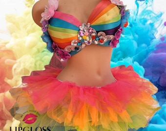 May include: A rainbow-colored bra with a jeweled center piece and flower embellishments, paired with a rainbow tulle skirt. The text "Be Sexy. Stand Out." is visible on the skirt.