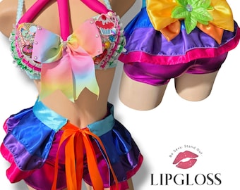 May include: A two-piece costume set featuring a decorated bra top with a large rainbow bow, cupcake and donut embellishments, and a halter strap. The matching skirt and shorts set includes layered satin ruffles in blue, purple, and pink.