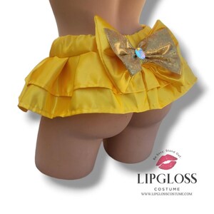 May include: A yellow ruffled costume bottom with a large gold bow and a central iridescent gem. The costume is made of a shiny fabric and is designed to be worn on the lower body. The costume has the text "LIPGLOSS COSTUME" at the bottom.