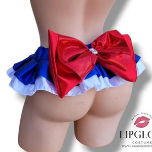 May include: A costume piece featuring a red bow and a ruffled skirt in blue, white, and red. The bow has a shiny, reflective surface. The skirt has a layered design with a white ruffled edge. The costume is designed to be worn around the waist.