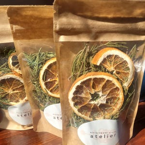 May include: Three brown kraft paper pouches with clear windows containing dried orange slices and green pine needles. Each pouch features a white oval label printed with the text bittersweet creek atelier in black lowercase letters.