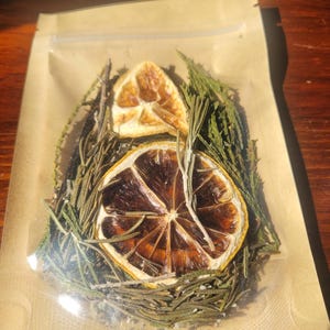 Puede incluir: Una bolsa transparente y resellable que contiene una mezcla de elementos botánicos secos, incluyendo dos rodajas de cítricos deshidratados, ramas de romero verde y agujas de coníferas, dispuestas dentro del envase de plástico.