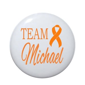 May include: White button with the text "TEAM Michael" in orange lettering and an orange ribbon.