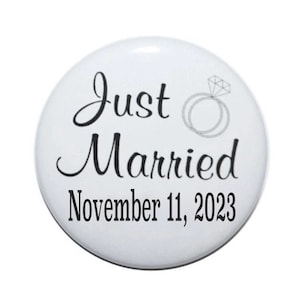May include: White button with black text that reads "Just Married" and "November 11, 2023". A simple ring with a diamond is above the text.