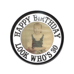 May include: A black and white round button with the text "Happy Birthday Look Who's 30" in a circle around a photo of a young girl.