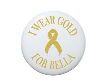 May include: White button with the text "I wear gold for Bella" in gold lettering. A gold ribbon is in the center of the button.