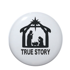 May include: A white, round button with a black silhouette of a nativity scene, including a star, stable, and figures. The text "TRUE STORY" is printed in black below the scene. The button has a glossy finish.
