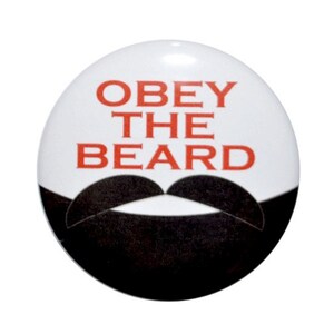 May include: A white button with the text "OBEY THE BEARD" in red letters and a black mustache underneath.