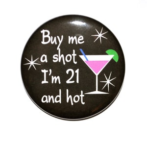 May include: Black button with white text that reads "Buy me a shot I'm 21 and hot" with a pink martini glass and a lime wedge on a black background.