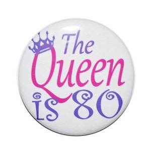 May include: A white button with a purple crown, pink text that says "The Queen is 80" and purple swirls.