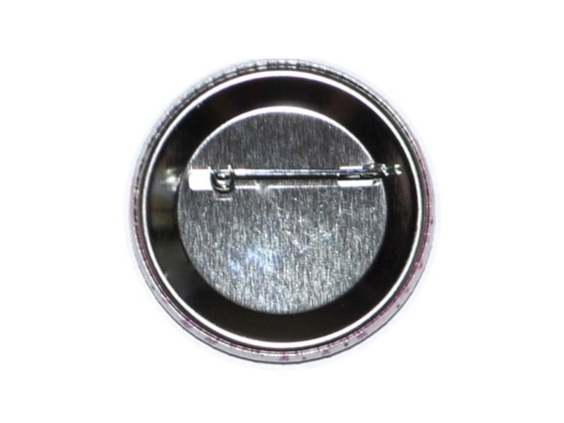 May include: The back of a round button with a silver metal backing and a pin on the back.