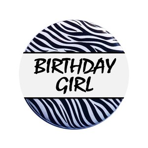 May include: A round button featuring a black and white zebra print design. The button has a white band with the text "BIRTHDAY GIRL" in black.