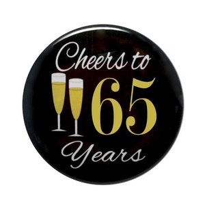 May include: Black button with white text that reads "Cheers to 65 Years" and two gold champagne glasses.