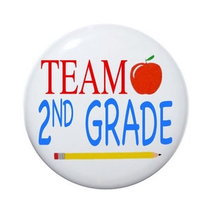 May include: A white button with the text "TEAM 2ND GRADE" in red and blue lettering. A red apple with a green stem and leaf is to the right of the word "TEAM". A yellow pencil with a red eraser is at the bottom of the button.
