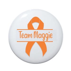 May include: White button with an orange ribbon and the text "Team Maggie" in orange.