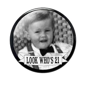 May include: A black and white circular button with a vintage photo of a young person wearing a bow tie and suspenders. The banner at the bottom reads "LOOK WHO'S 21".